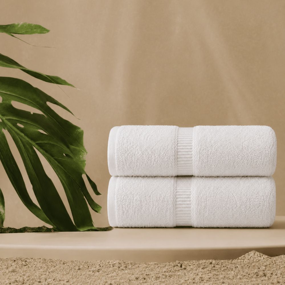 Two white CH Series premium bath towels stacked in a warm spa setting with a tropical leaf and elegant textured centre bands.
