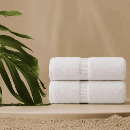 Two white CH Series premium bath towels stacked in a warm spa setting with a tropical leaf and elegant textured centre bands.