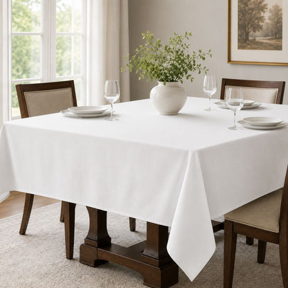 Classic solid white tablecloth on a rectangular dining table with white dinnerware, wine glasses and a simple greenery centerpiece.