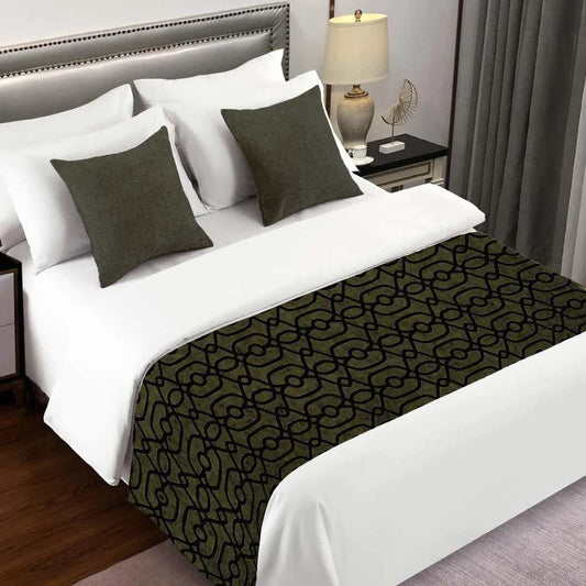 A hotel room with a bed next to a lamp on a nightstand. Has white sheets with two decorative pillows in a deep moss green colour. Towards the end of the bed there is a bed scarf/ runner in a deep moss green colour with black details thought out the fabric.