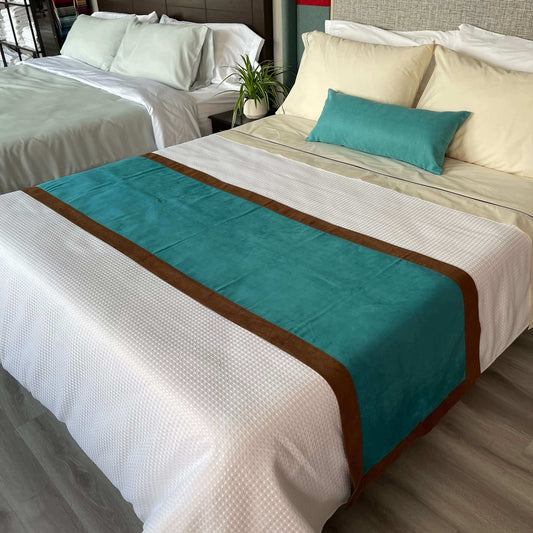 A bed with white and yellow sheets with decorative pillows near the headboard.  Towards the end of the bed there is a custom reversible bed scarf/ runner  that is brown and turquoise 